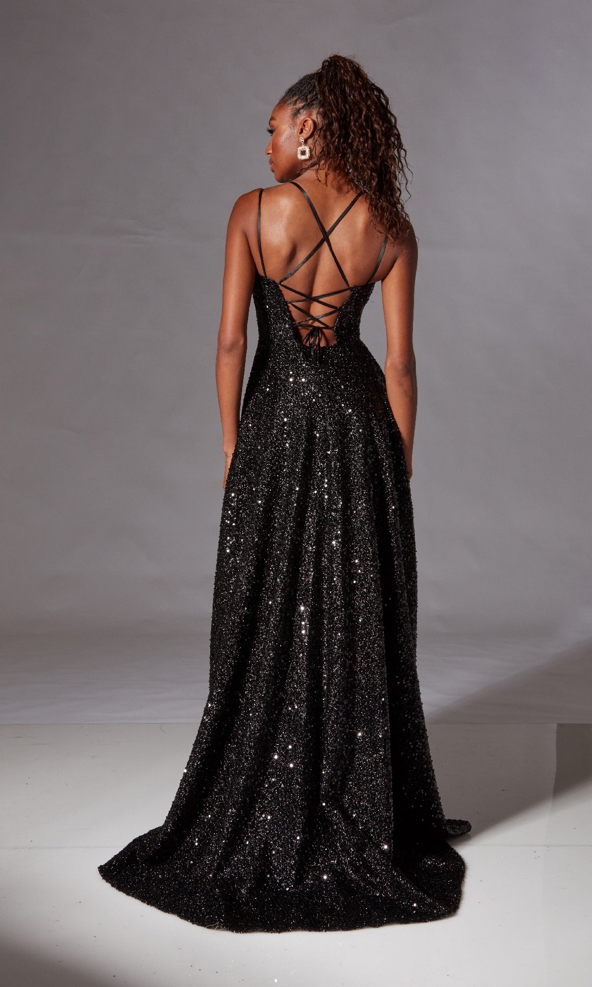 A woman stands facing away against a plain backdrop, wearing the Aerie Couture 273 Long Black Glitter Prom Dress with a crisscross lace-up open back.